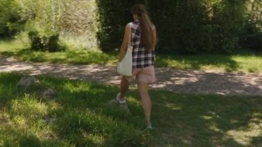 Güneş lekeli çakıl yolu üzerinde yürüyen bir kadın, Lush Green Park 'ta, bir çanta tutarken geriye bakıyor.