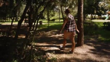 Güneş ışığı yaprakların arasından süzülürken, Ağaçlar ve banklarla çevrili, Gölgeli Orman yolunda yürüyen bir kadın..