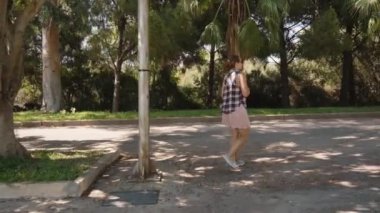 Kadın, uzun ağaçlarla çevrili bir parkta, güneşli bir yolda yürüyor ve kaldırımlarda gölgeler bırakıyor..