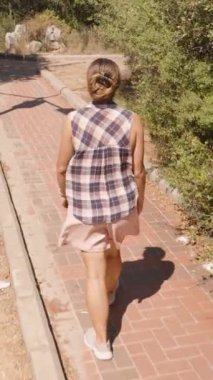 Dikey video. Kırmızı Tuğla Yolu 'nda yürüyen bir kadın Sunlit Çalıları ve Ağaçları ile çevrili Yoldaki Çatal' a yaklaşıyor..