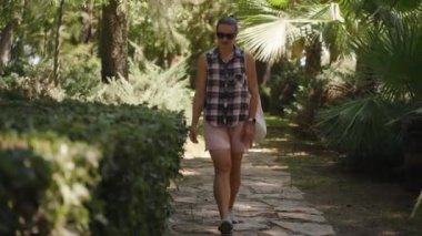 Güneş gözlüklü ve ekose gömlekli, Tropikal ağaçlar ve çalılarla çevrili bir kadın..