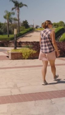 Dikey video. Kadın, bir şehir parkında Parlak Günışığı 'nın altında düzgünce süslenmiş çalılar ve merdivenlerle Paved Plaza' da geziniyor..