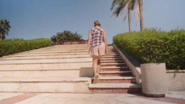 Kadın, açık mavi gökyüzünün altında Yeşillik ve Palmiye ağaçlarıyla çevrili bir dizi taş basamağa tırmanıyor..