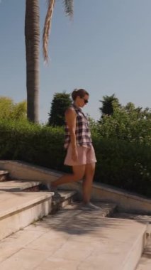 Dikey video. Geniş Taş Merdiven 'de yürüyen bir kadın. Kenarlarında Yeşil Çitler var. Açık Gökyüzünün altındaki bir parkla çevrili..