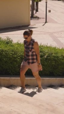 Dikey video. Lush Green Hedges 'in yanında bir taş merdivene tırmanan kadının adımları kaldırıma gölge düşürüyor..