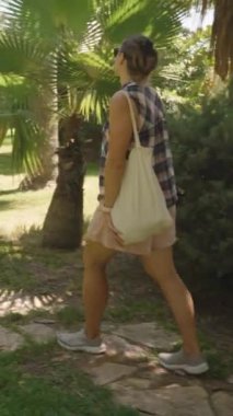 Dikey video. Güneş Işığı Bahçesi 'nde yürüyen bir kadın. Etrafı bereketli tropikal bitkilerle çevrili. Çimenlere yumuşak gölgeler serpiştiriyor..