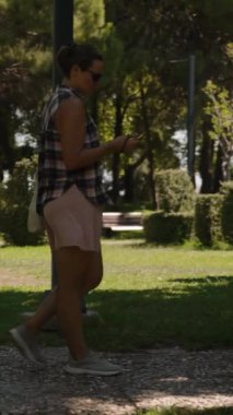 Dikey video. Kadın gölgeli bir parkta yürüyor, telefonuna odaklanmış, ağaçlar ve güneş lekeli otlarla çevrili. Yavaş çekim.