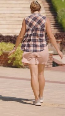 Dikey video. Ekose gömlekli ve pembe şortlu bir kadın güneşli bir günde Lush Garden ve Cascading Fountain 'ın önünden geçiyor. Yavaş çekim.