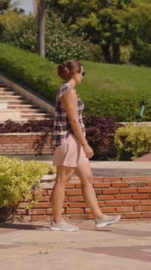 Dikey video. Bir Sunlit Yolu 'nda Neşeli Dengeli Kolları Olan Kadın, Ağaçlar ve Çalılar' ın yanında günlük kıyafetler giyiyor. Yavaş çekim.