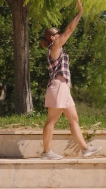 Dikey video. Ekose gömlekli kadın ve pembe şortlu kadın Sunlit Steps 'te dikilirken Yeşil Ağaç Dallarına doğru uzanıyor. Yavaş çekim.