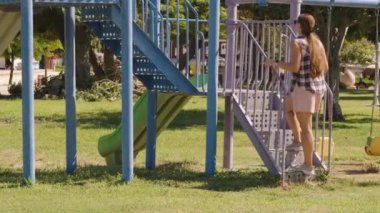 Ekose tişörtlü ve pembe şortlu bir kadın güneşli bir günde Yeşil Ağaçlar 'ın altındaki mavi çocuk parkı merdivenlerine tırmanıyor. Yavaş çekim.