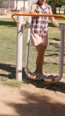 Dikey video. Ekose gömlekli ve pembe şortlu kadın açık hava altında Yeşil Park 'ta egzersiz ekipmanı kullanıyor. Yavaş çekim.