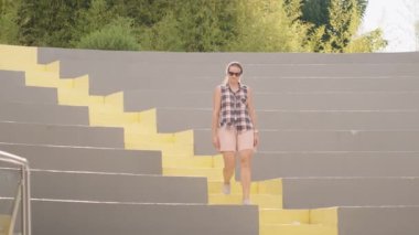 Ekose gömlekli bir kadın yeşil bambuyla çevrili, modern bir açık hava amfitiyatrosunda sarı basamaklı koltuklarda yürüyor. Yavaş çekim.