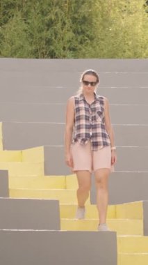 Dikey video. Ekose gömlekli bir kadın yeşil bambuyla çevrili, modern bir açık hava amfitiyatrosunda sarı basamaklı koltuklarda yürüyor. Yavaş çekim.
