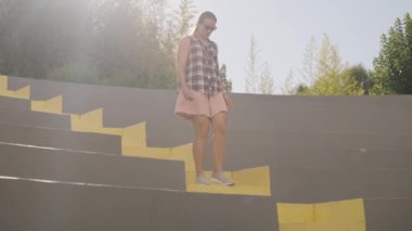 Bir kadın açık hava amfitiyatrosunda özenle sarı basamaklarda yürür, yumuşak güneş ışığıyla yıkanır. Yavaş çekim.