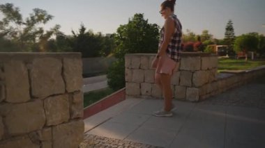 Gündelik giysiler içindeki kadın, taştan duvarlarla çevrili güneşli bir alana doğru iniyor. Ağaçlar ve yumuşak akşam ışığı sakin bir atmosfer yaratıyor..
