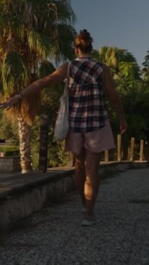 Dikey video. Kareli Gömlek ve Pembe Şortlu bir kadın, ağaçlarla kaplı bir patikada yürüyor. Huzurlu bir akşam boyunca gölgeleri yere doğru uzanıyor..