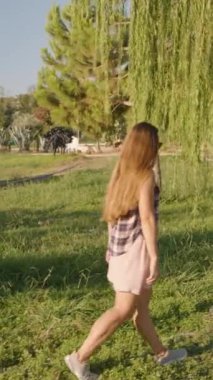 Dikey video. Etrafı yemyeşil otlarla çevrili, Ağlayan Söğüt 'ün gölgesinin altında gezinen bir kadın..