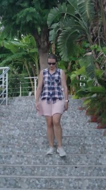 Dikey video. Tropikal Yapraklar ve Modern Bina 'yla kaplı Büyük Taş Merdivenden İnen Kadın..