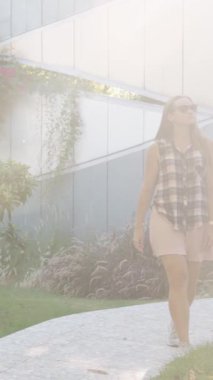 Dikey video. Ekoseli Gömlekli Kadın Gün Işığı Gibi Yürüyor, Çevresi Yeşillik ve Arka planda Modern Bir Bina ile çevrili.