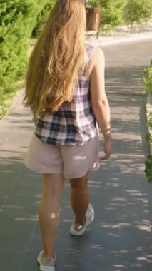 Dikey video. Ekose gömlekli bir kadın, Gölgeli bir yol boyunca yürüyor. Etrafı yeşil çalılar ve ağaçlarla çevrili. Gün ışığının yumuşak gölgeleri var..