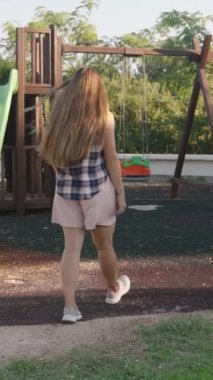 Dikey video. Bir kadın oyun parkına doğru yürüyor, bir salıncak setine yaklaşıyor, arkasında uzun saçlar uçuşuyor..