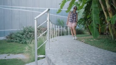 Ekose gömlekli kadın kavisli taş basamaklarda yürüyor, etrafı yemyeşil ağaçlar ve metal parmaklıklarla çevrili..