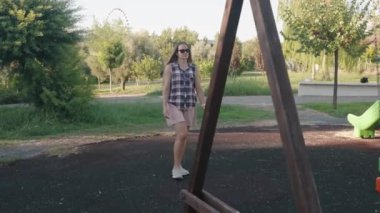 Ekoseli Gömlekli Kadın Çocuk Bahçesinde Yürüyor Sallanıyor, Arka planda Huzurlu Park Ayarları Var.