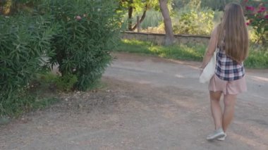 Geniş yeşil bir çalılığın yanındaki parkta yürüyen, kolsuz bir gömlek ve pembe etek giyen, omzunda beyaz bir çantayla, güneşli günün tadını çıkaran bir kadın..