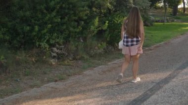 Park yolunda yürüyen, günlük kıyafetli bir kadın yeşil kuşakların tadını çıkarıyor, yerde uzun gölgeler bırakan güneş..