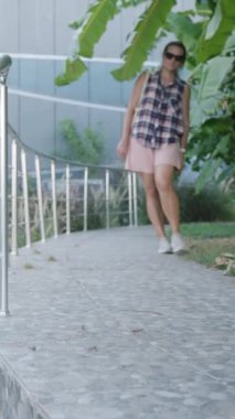 Dikey video. Ekose gömlekli kadın kavisli taş basamaklarda yürüyor, etrafı yemyeşil ağaçlar ve metal parmaklıklarla çevrili..