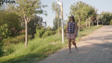 Ağaçlarla çevrili bir yolda yürürken, günlük giysiler içindeki bir kadın huzurlu bir anın tadını çıkarırken, gözleri manzarada aşağı doğru odaklanmış..