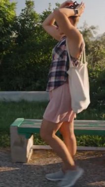 Dikey video. Güneşli bir parkta yürüyen, saçlarını düzelten, yemyeşil ve ılık akşam ışığında yıkanan bir kadın..