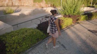 Kareli Gömlekli Kadın ve Pembe Şortlu Kadın güneşli bir yolda yürüyor, Arkası kameraya dönük, gölgeler ilerlerken beyaz bir çanta taşıyor.