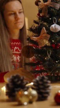Dikey video. Kırmızı kazaklı bir kadın Noel ağacını dikkatlice inceliyor ve her şeyin mükemmel olmasını sağlıyor..