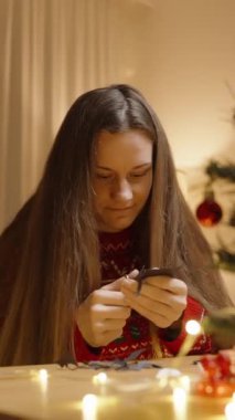 Dikey video. Kırmızı Süveterli Bir Kadın, Tatil Süslemeleri ve Sıcak ve Şenlikli Atmosfer Oluşturan Bir Masada El Yapmaya Odaklanıyor.