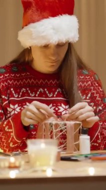 Dikey video. Kırmızı Süveterli Kadın ve Noel Şapkalı Kadın Sıcak Bir Evde Şölen Dekoru ile süslenmiş bir masada Noel süslemesi yapmaya odaklanıyor.