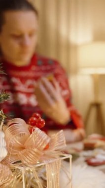 Dikey video. Kırmızı kazaklı bir kadın, Noel Ağacı Kurabiyesini Gösteriyor, Güzel Aydınlatılmış Noel Ağacının yanında.