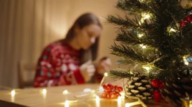 Işıklar ve süslemelerle süslenmiş bir Noel Ağacı Odakta, Arkaplanda El Sanatları Üzerinde Çalışan Bir Kadın.