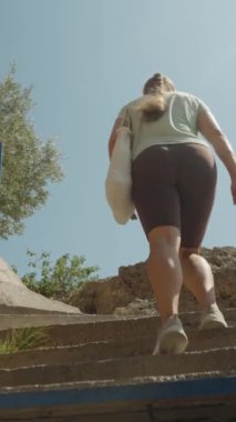 Dikey video. Taş Merdivenlerin Üzerindeki Kadın, Yeşillik ve Kayalık Arazi İki Tarafta Mavi Raylar Yolu İşaretliyor, Açık Gökyüzü.
