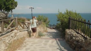 Seaside Stroll, Stone Yolu 'nda Yürüyen Kadın, Sağında Okyanus, Solunda Ahşap Çit ve Yeşillik, Açık Gökyüzü.