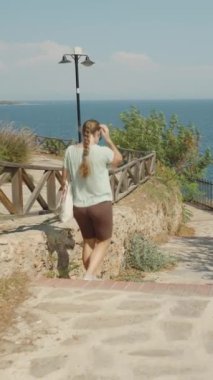 Dikey video. Seaside Stroll, Stone Yolu 'nda Yürüyen Kadın, Sağında Okyanus, Solunda Ahşap Çit ve Yeşillik, Açık Gökyüzü.