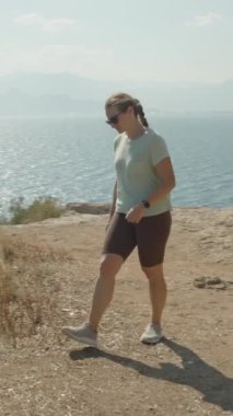 Dikey video. Uçurumun Kıyısında Duran Kadın, Aşağıdaki Parıldayan Okyanusa bakıyor, Etrafı Rocky Arazisi ile çevrili, Uzakta Dağlar.