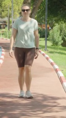 Dikey video. Ön Manzara, Kırmızı Yol 'da Yürüyen Kadın, Çevresi Yeşil ve Ağaçlarla çevrili, Solunda Mavi Bisiklet Yolu, Tepesinde Güneş Işığı.