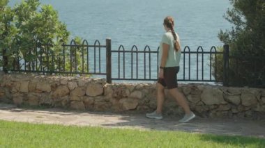 Okyanusa Yakın Yürüyen Kadın, Altında Taştan Yol, Yanda Yeşil Çimen, Kıyıda Demir Çit, Açık Gök ve Sakin Deniz.