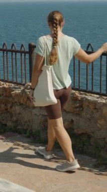 Dikey video. Sarmal Taş Yoldan Aşağı Yürüyen Kadın, Okyanusa Bakan, Çantayı Tutan Demir Çit ve Taş Duvarlar, Parlak Gün.