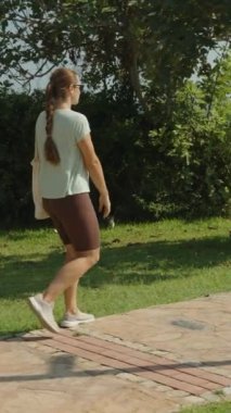 Dikey video. Okyanus kenarındaki Taş Yolda Yürüyen Kadın, Piknik Masası ve Yeşillik 'ten Geçen, Açık Gökyüzü ve Arka planda Sakin Deniz' le Parlak Gün.