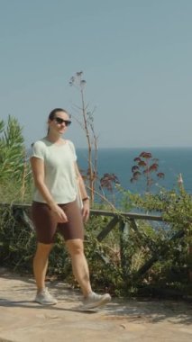 Dikey video. Side View, Deniz kenarında gezinen kadın, yeşillikle çevrili, Piknik masasını geçen, arka planda okyanusu geçen..