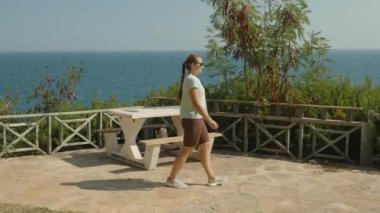 Taş Patio Bölgesinde Yürüyen Kadın, Okyanusa Bakan, Piknik Masası Yakın, Yeşillik ve Açık Gökyüzü Huzur Oluşturuyor.