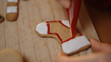 Hand Outlines a Stocking Cookie With Red Icing Over White Frosting, While Decorated Christmas-Themed Cookies Lie on the Wooden Board. clipart